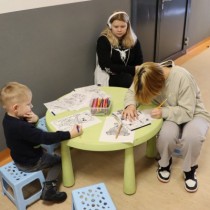 Powiększ zdjęcie Kącik plastyczny. Trójka dzieci siedzi wokół okrągłego stolika i maluje kredkami wydrukowane obrazki.