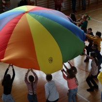 Powiększ zdjęcie Dzieci podczas zabawy z kolorową chustą animacyjną. Uczestnicy trzymają wielobarwną tkaninę i podrzucają ją do góry.