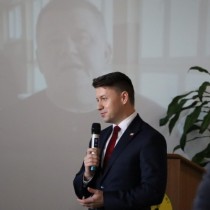 Powiększ zdjęcie Poseł Bartłomiej Dorywalski trzyma mikrofon, przemawia do zebranych uczestników uroczystości z okazji 37. rocznicy protestu o krzyże w Zespole Szkół Zawodowych we Włoszczowie.
