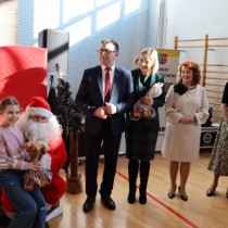 Powiększ zdjęcie Przy kąciku świetego Mikołaja. Mikołaj wręcza prezent dziewczynce, obok stoją starosta Dariusz Czechowski, dyrektor PCKR Teresa Chudy - trzyma paczkę, członek Zarządu Małgorzata Gusta, dyrektor PCPR Ilona Nowak.