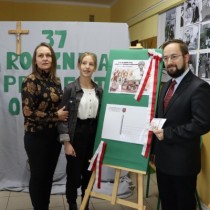 Powiększ zdjęcie W środku sztaluga z makietą kartki jubileuszowej. Po prawej Członek Zarządu Powiatu Włoszczowskiego i Dyrektor Szkoły Podstawowej w Olesznie Rafał Pacanowski po lewej laureatka z mamą..