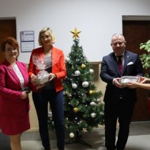 Powiększ zdjęcie W środku choinka udekorowana białymi i złotymi dekoracjami. Od lewej stoją: Członek Zarządu Małgorzata Gusta odbiera piernik od Renaty Dziedzic, Wicestarosta Włoszczowski Łukasz Karpiński odbiera piernik od Wioletty Hupy.