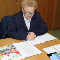 Powiększ zdjęcie Teresa Ostrowska siedzi, w ręce trzyma długopis nad otwartą czystą stronicą książki. Na stoliku po lewej stronie otwarta książka.