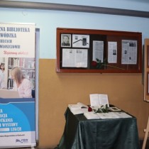 Powiększ zdjęcie Po lewej stronie stoi baner reklamowy Pedagogicznej Biblioteki Wojewódzkiej w Kielcach obok stolik z otwartymi książkami i czerwoną różą pośrodku. Nad stolikiem tablica poświęcona sylwetce Łukasza Uniejewskiego.
