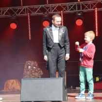Powiększ zdjęcie Na scenie po prawej stronie dziecko asystujące magikowi w czarnym fraku (z lewej).