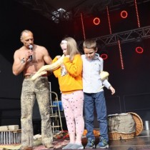 Powiększ zdjęcie Na scenie dwójka dzieci po prawej trzyma żółtego węża boa. Po lewej stronie stoi mężczyzna z mikrofonem.