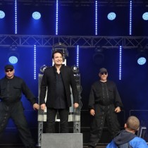 Powiększ zdjęcie Na scenie stoi trzech mężczyzn w czarnych ubraniach.