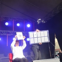 Powiększ zdjęcie Pokaz iluzji. Na scenie siedzi kobieta w białej kurtce trzymając nad głowa kartę, obok po prawej mężczyzna trzyma tablicę z talią kart.
