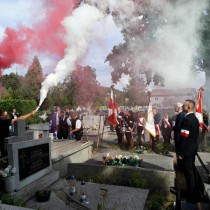 Powiększ zdjęcie Uczestnicy pogrzebu na cmentarzu. Na pierwszym planie płoną race w kolorze białym i czerwnonym.