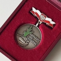 Powiększ zdjęcie Okrągły medal. W środku medalu jest umieszczona płaskorzeźba przedstawiająca dłonie trzymające garść ziemi, z której wyrasta roślina. Roślina jest pokryta zieloną emalią. Obok rośliny jest umieszczony napis 